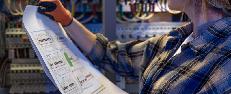 A woman electrician holds printed schematics and cross checks them against power modules and wiring in a cabinet. Blue lighting sets a careful mood of quality control and safe verification.
