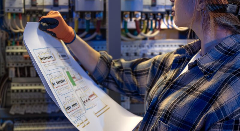 A woman electrician holds printed schematics and cross checks them against power modules and wiring in a cabinet. Blue lighting sets a careful mood of quality control and safe verification.
